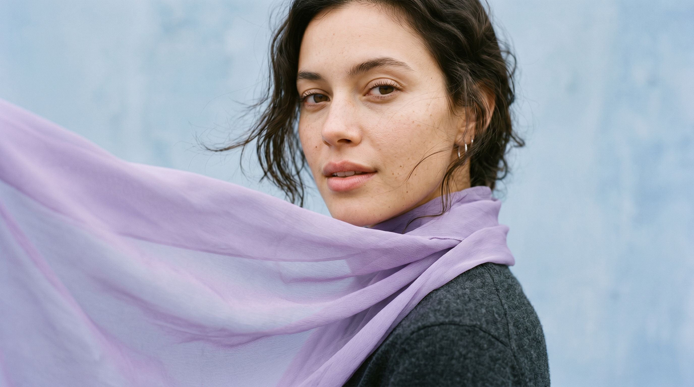AI-generated on-model fashion image: close-up portrait of a model in a grey wool coat and lilac silk scarf against a pale blue wall