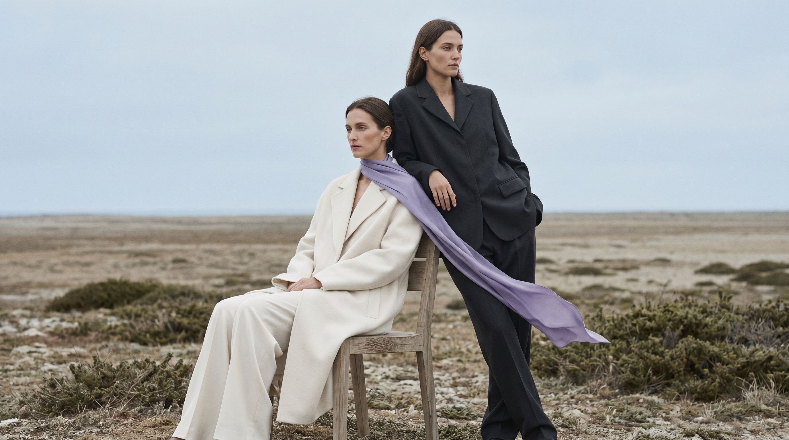 AI-generated on-model fashion image: two models on coastal scrubland, one seated in a cream wool coat and trousers, one standing in a charcoal tailored suit, joined by a lilac silk scarf